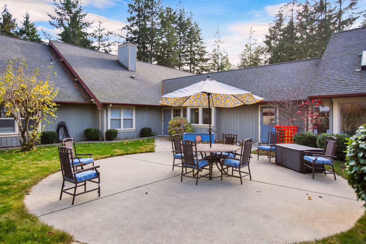 Outdoor patio area at Greenlake Senior Living Gig Harbor with a round table and six chairs under a large umbrella. The patio is surrounded by a building with gray siding and a shingled roof, with trees and shrubs in the background.