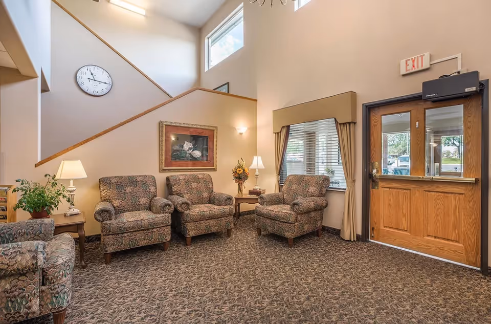 A cozy seating area in a senior living facility with four patterned armchairs arranged around two wooden side tables, each holding a lamp and a plant or flowers. The room has beige walls, a large clock on the wall, a framed floral painting, a window with beige curtains, and a wooden exit door with glass panels.