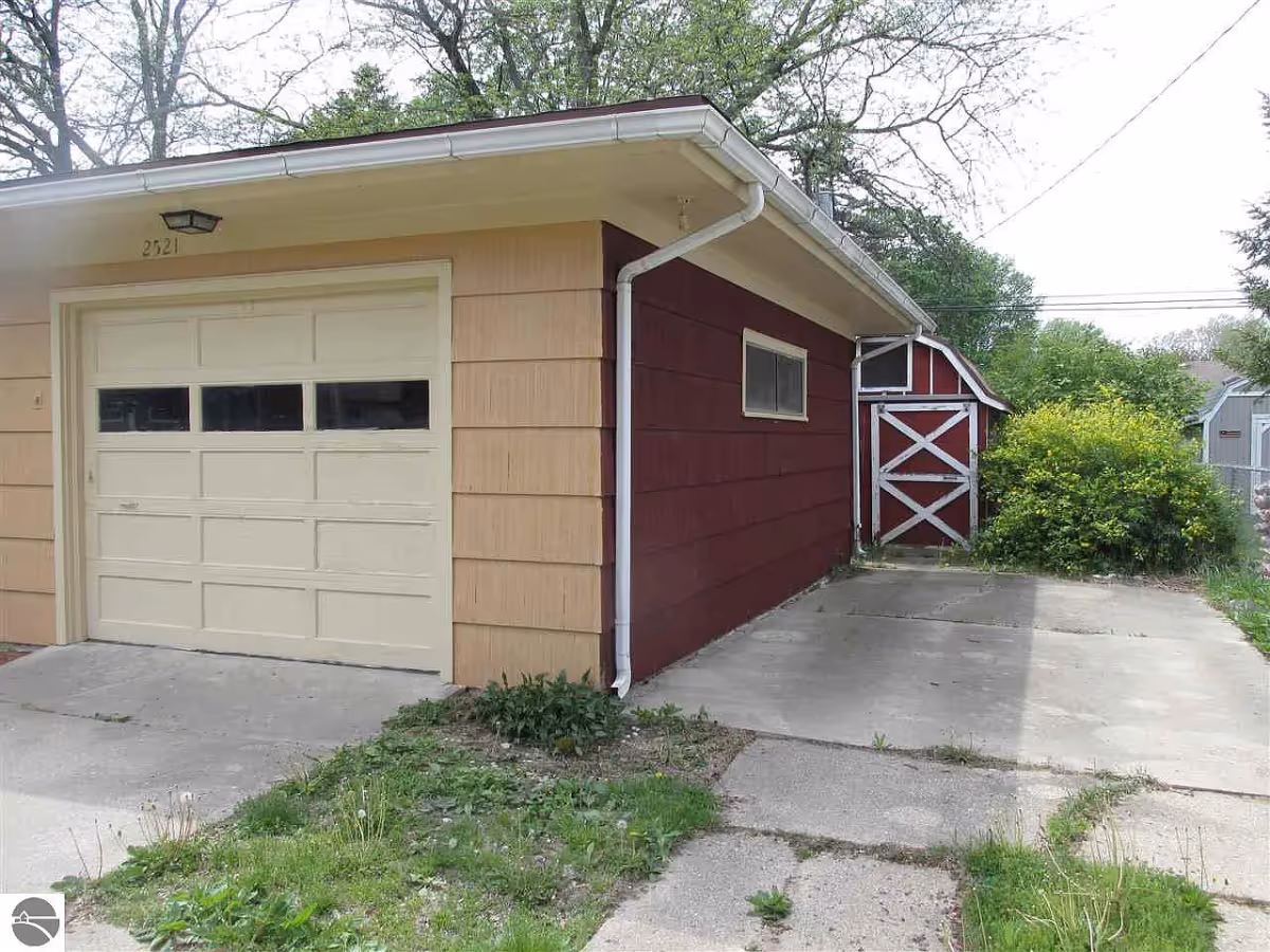 Driveway in front of a beige garage door on a house with red siding and a red-and-white shed at the back.