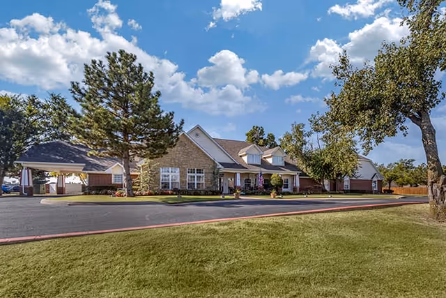 Exterior view of a single-story senior living facility building with a stone facade, multiple windows, and a covered entrance. The building is surrounded by a well-maintained lawn, trees, and a paved driveway under a partly cloudy blue sky.