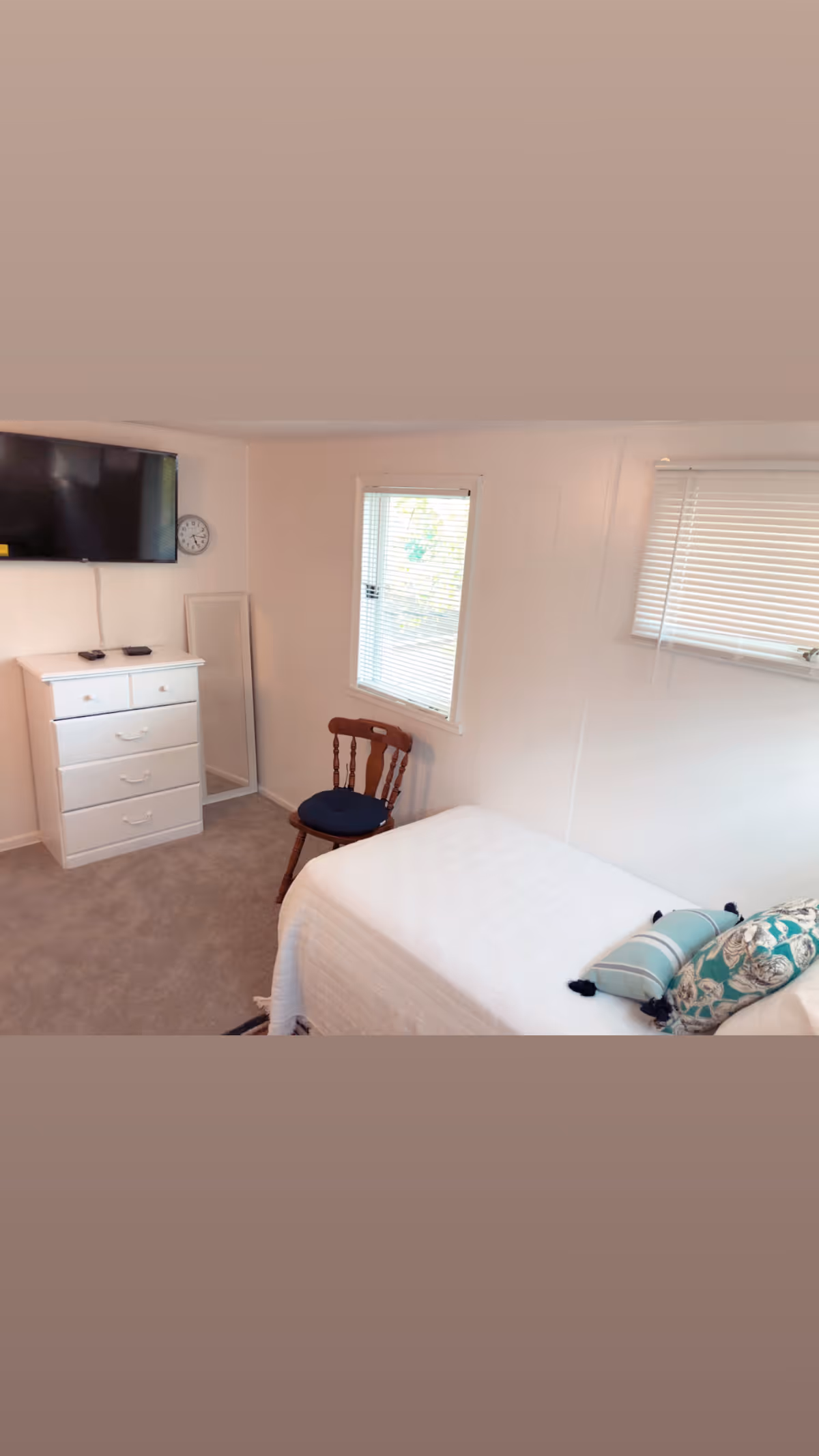 A small bedroom with a single bed covered in a white bedspread and decorative pillows. There is a wooden chair with a dark cushion next to a window with blinds. A white chest of drawers with a flat-screen TV mounted above it and a wall clock is visible. The room has light-colored walls and carpeted floor.