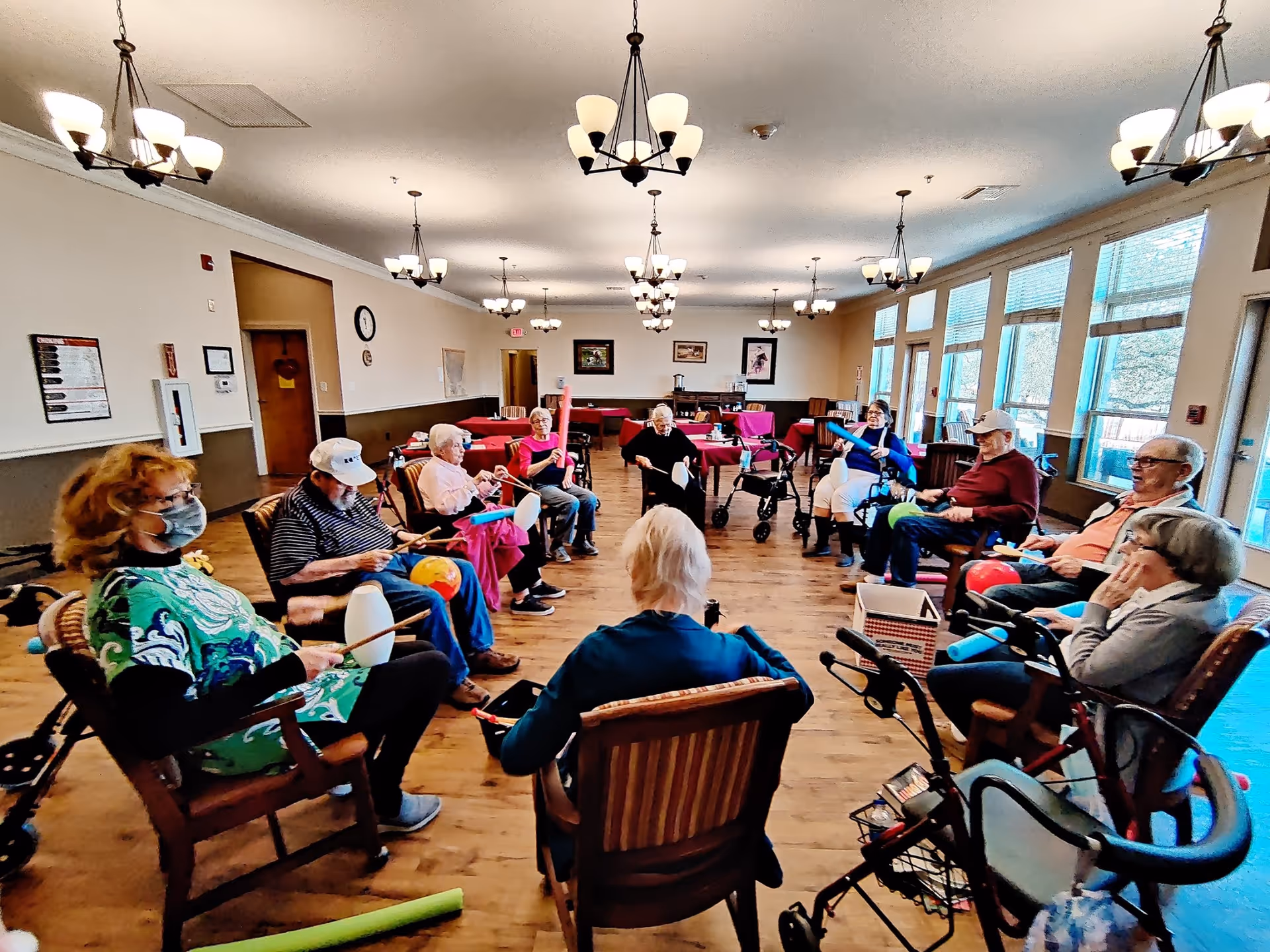 A group of elderly people sitting in a circle in a large, well-lit room with wooden floors and multiple chandeliers. They are engaged in an activity using colorful drumsticks and percussion instruments. Some have walkers nearby, and large windows line one side of the room.