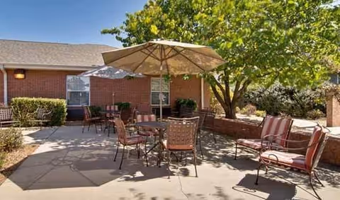 Outdoor patio area with metal tables and chairs, some with cushions, under a large beige umbrella. The patio is surrounded by a brick building and greenery including a tree providing shade.