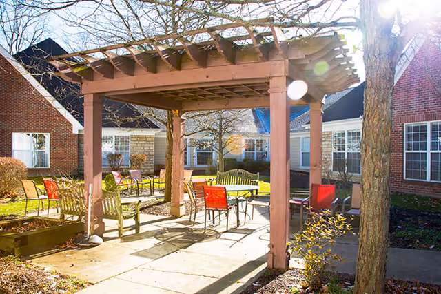 Outdoor patio area with a wooden pergola providing partial shade over several chairs and tables. The patio is surrounded by a brick building with multiple windows, and there are leafless trees and some shrubs around the area. Sunlight creates lens flare effects in the image.