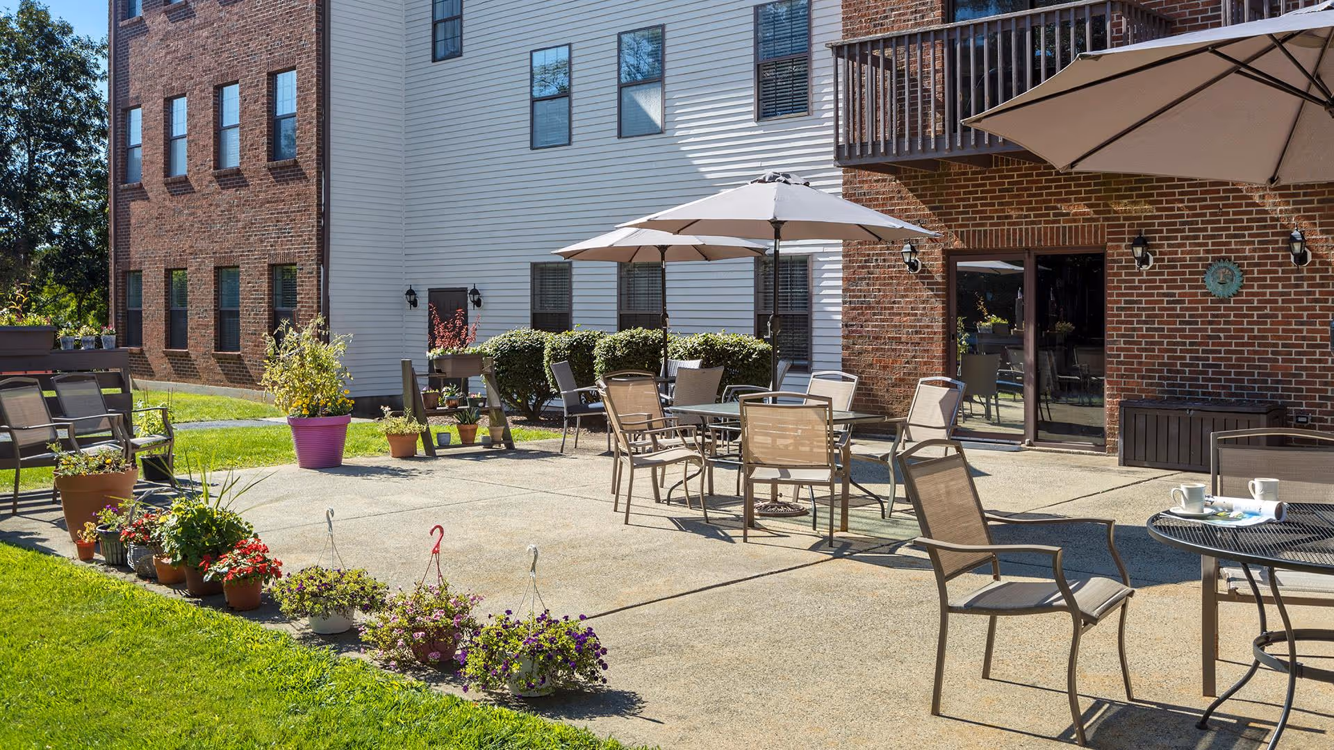 Outdoor patio area at Cedar Woods facility with multiple tables and chairs under large umbrellas, surrounded by potted plants and flowers. The patio is adjacent to a brick and white siding building with windows and a glass door.