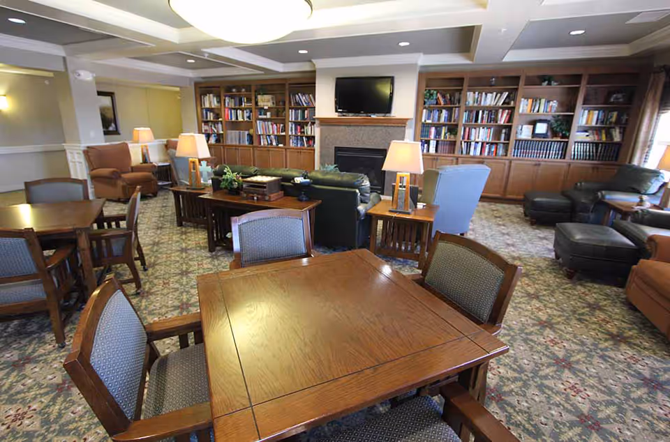 Spacious senior living common room with wooden tables and chairs, armchairs and sofas, bookshelves, lamps, and a TV above a fireplace.