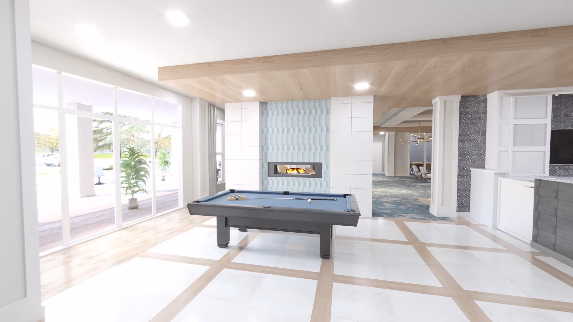 A modern indoor recreational area with a pool table in the center, a contemporary fireplace with blue textured tiles behind it, large floor-to-ceiling windows letting in natural light, and a view of an outdoor patio with plants.
