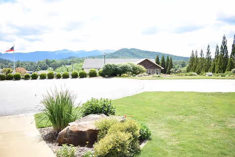 A landscaped outdoor area with green grass, shrubs, and a large rock in the foreground. A paved driveway curves around the greenery. In the background, there is a single-story building with a sloped roof, surrounded by trees and bushes. Mountains and a partly cloudy sky are visible in the distance.