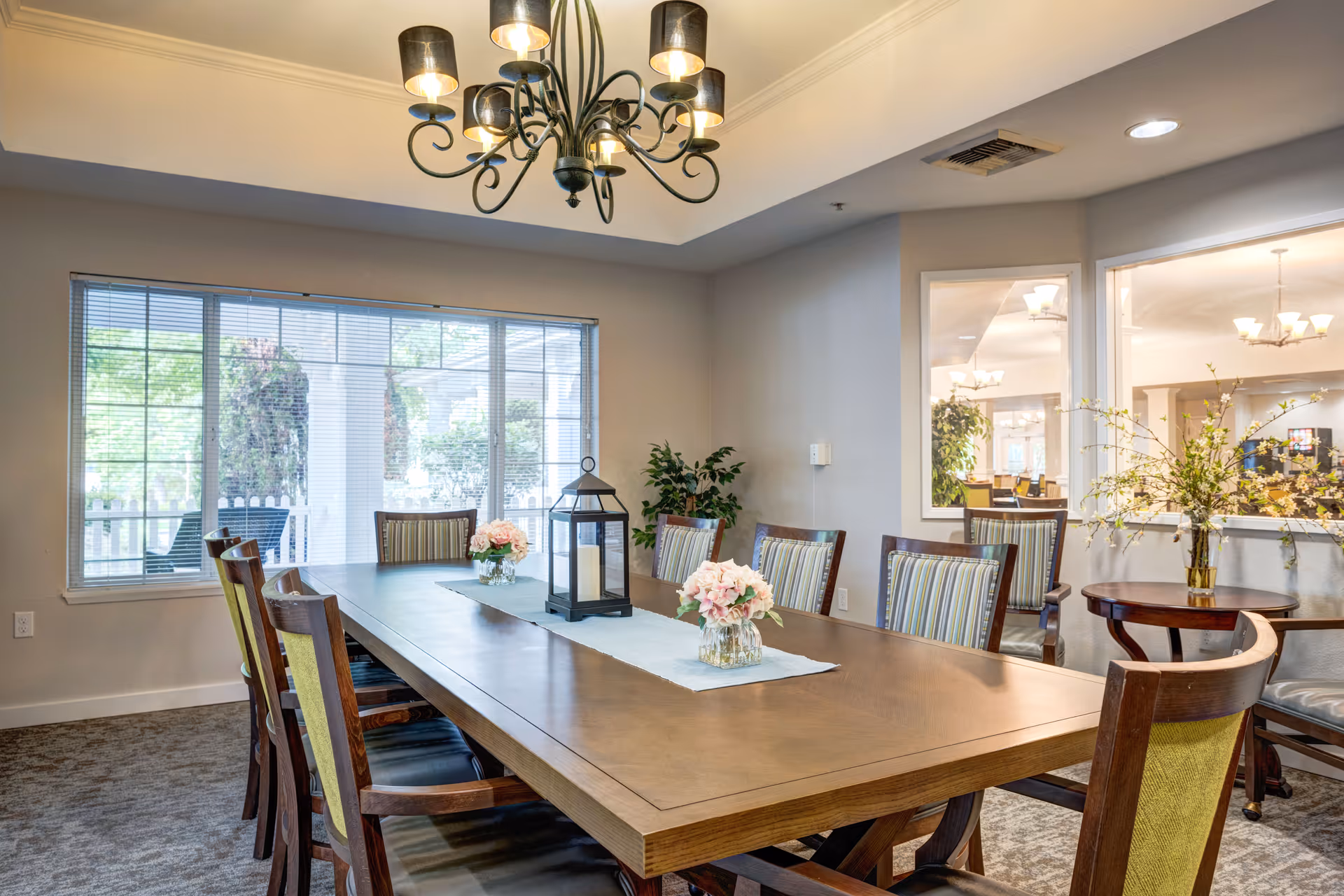 A well-lit dining room with a long wooden table surrounded by chairs with striped cushions. The table is decorated with a light blue runner, two vases of pink flowers, and a black lantern centerpiece. A large window with blinds lets in natural light, and a chandelier with multiple lights hangs from the ceiling. There is a small round table with a vase of flowers in the corner, and a large window looking into another room.