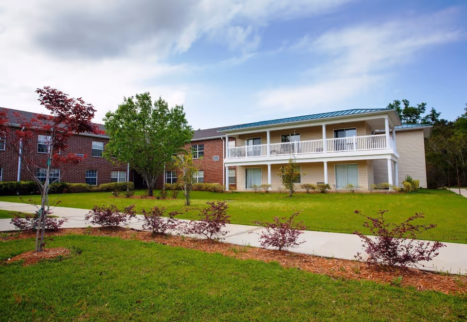 Exterior view of a senior living facility named Hatton House featuring a two-story building with a brick section and a beige siding section with a balcony. The foreground shows a well-maintained lawn, small trees, and a concrete walkway under a partly cloudy sky.