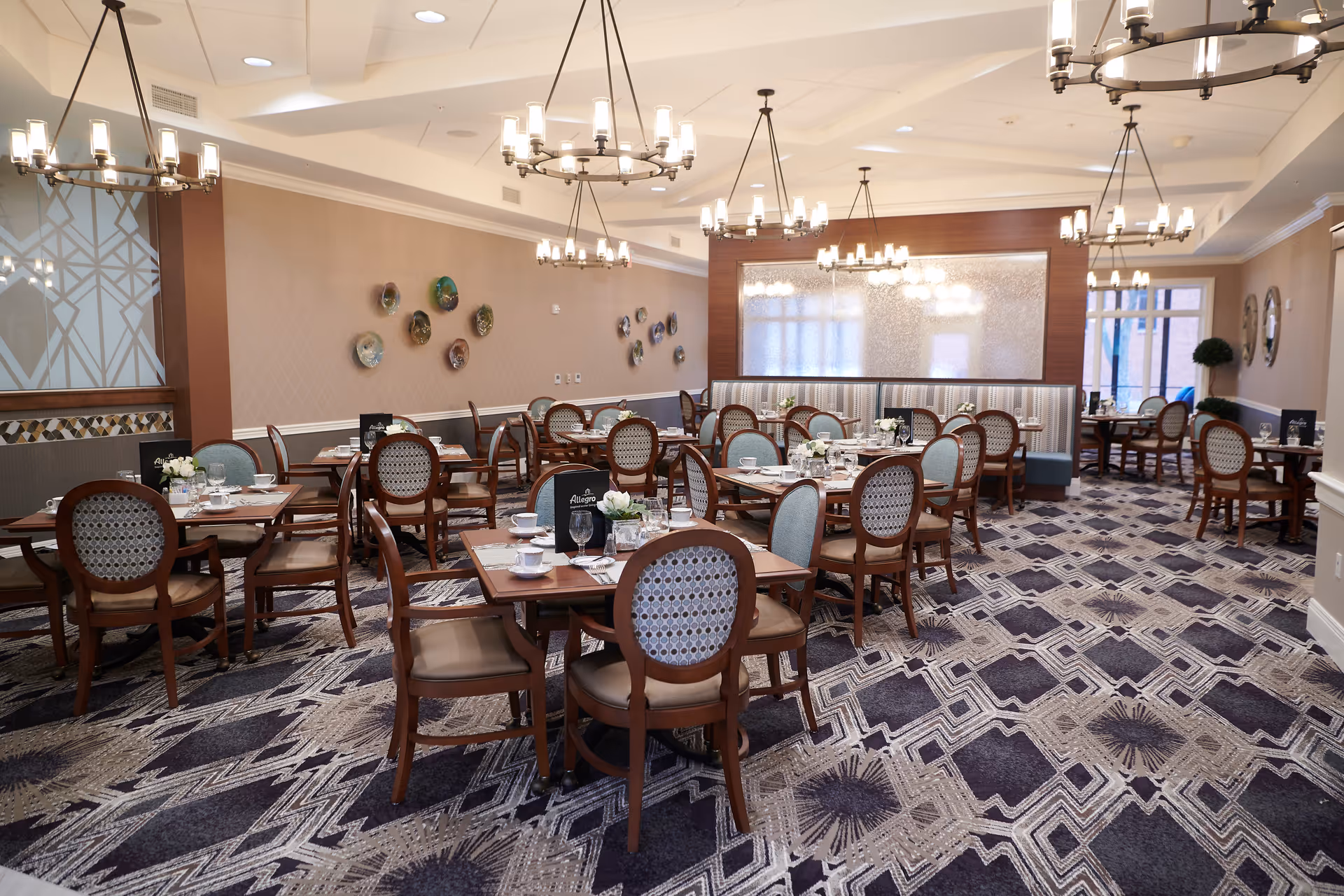 A spacious and elegantly decorated dining room with multiple wooden tables and chairs arranged neatly. The room features patterned carpet flooring, chandeliers hanging from the ceiling, decorative plates on the wall, and place settings with cups, glasses, and menus on the tables.