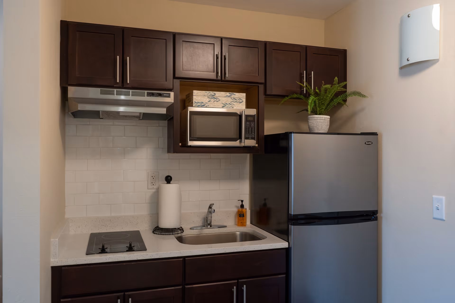 A compact kitchen area with dark wooden cabinets, a small stainless steel refrigerator, a microwave, a sink with a faucet, a two-burner electric stovetop, a paper towel holder, a soap dispenser, and a potted plant on top of the refrigerator. The backsplash is white subway tile and the walls are light-colored.
