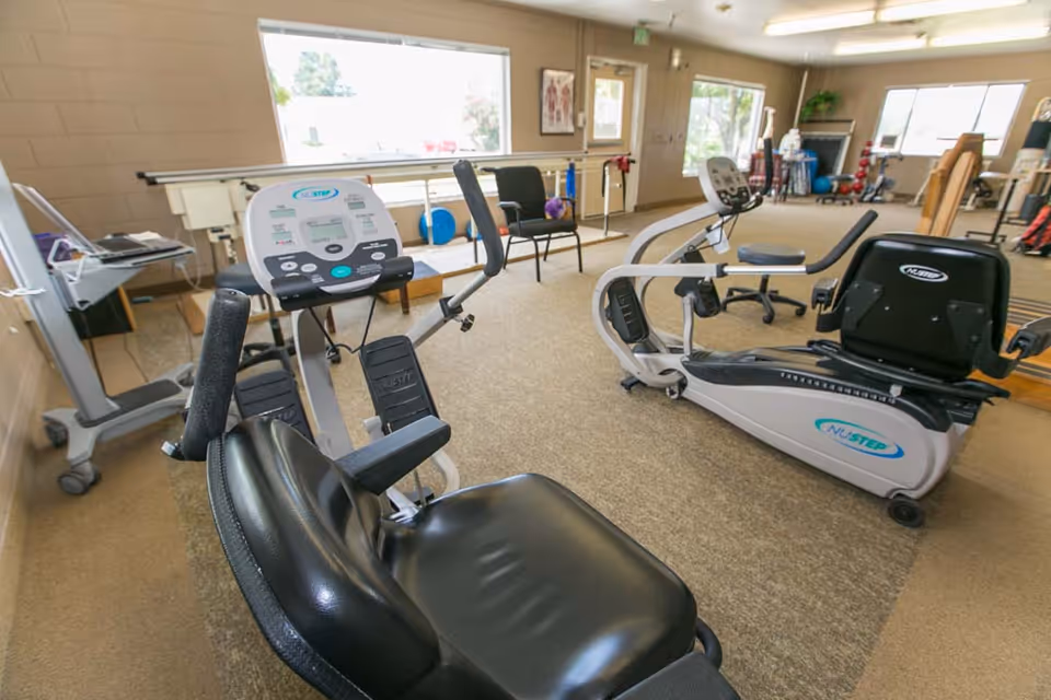 A rehabilitation exercise room with two NuStep recumbent cross trainers, chairs, exercise equipment, and large windows letting in natural light.