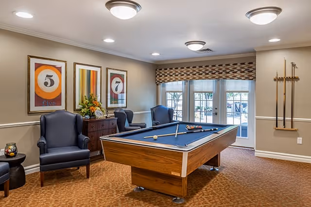 A well-lit game room with a pool table in the center, surrounded by four blue armchairs. The walls are decorated with three framed colorful prints, and there is a wooden cabinet with a flower arrangement on top. A set of pool cues is mounted on the wall near a set of glass double doors with patterned valances, leading outside.