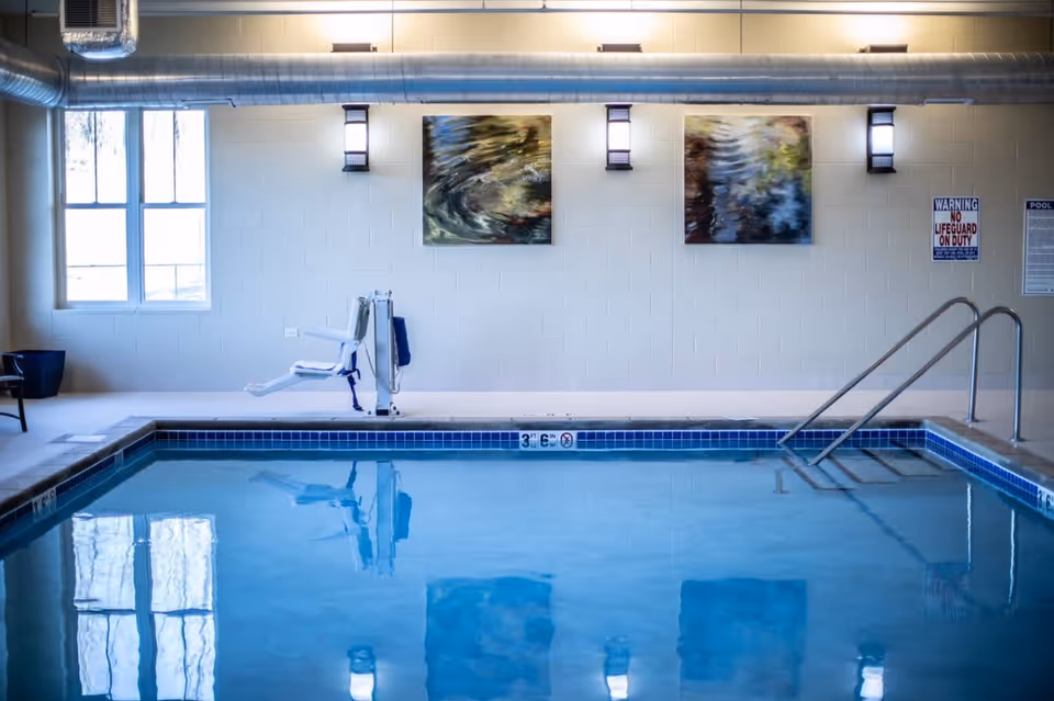 Indoor swimming pool with clear blue water, a pool lift chair on the left side, metal handrails on the right side, two abstract paintings on the wall, and a window letting in natural light.