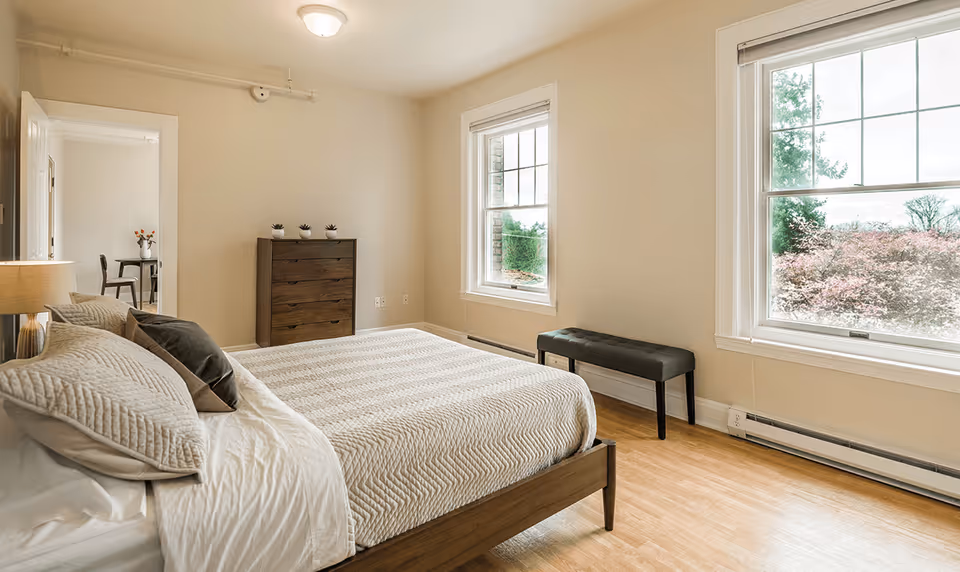 A bright bedroom with a neatly made bed featuring beige and brown pillows and a textured beige bedspread. The room has light-colored walls and wooden flooring. There are two large windows letting in natural light and showing greenery outside. A dark wooden dresser with three small potted plants on top is against one wall, and a black cushioned bench is placed under one window. An open door reveals a glimpse of another room with a table and chairs.