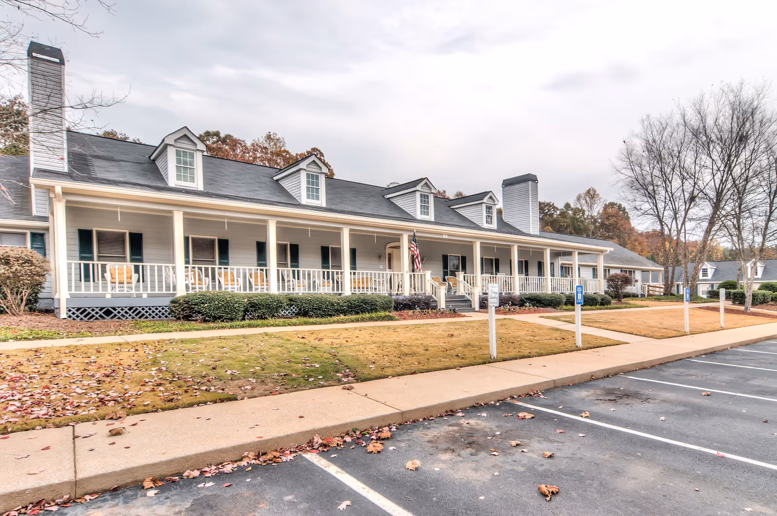 Long single-story senior living building with a covered front porch, white railings, an American flag, and a parking lot in front.