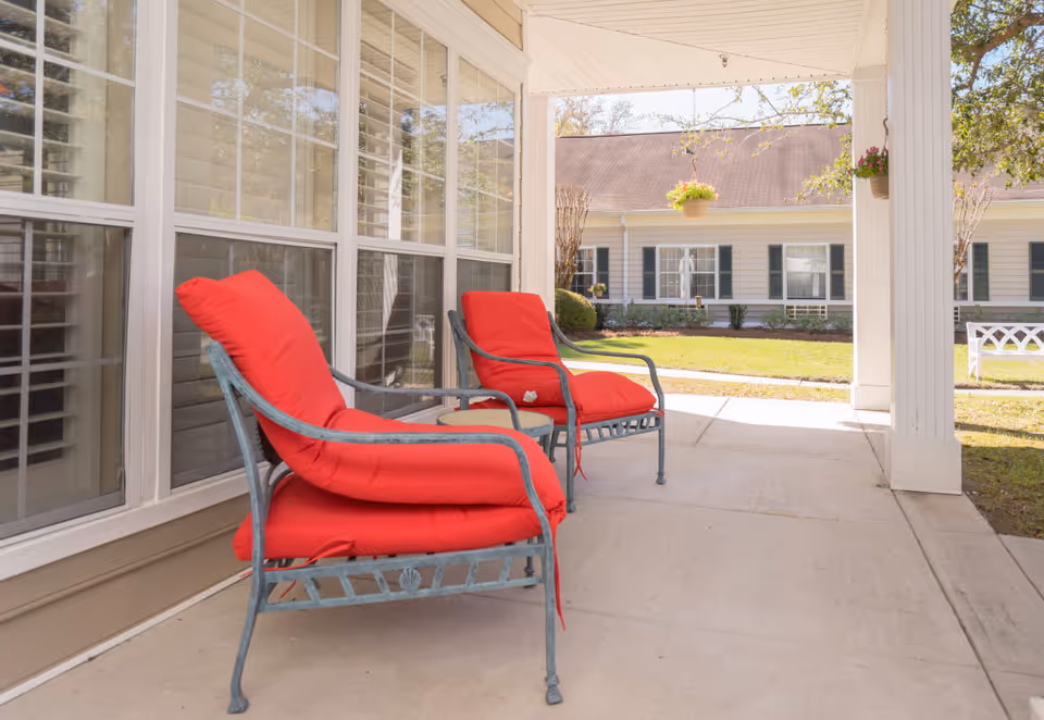 Two metal patio chairs with bright red cushions sit on a covered porch area outside a building with large windows. In the background, there is a lawn with shrubs, a hanging flower pot, and another building with white siding and dark shutters.