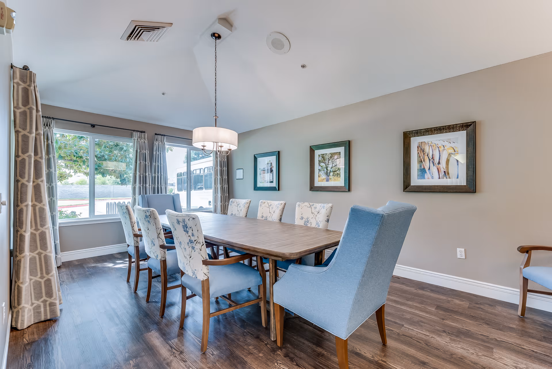 A bright dining room with a wooden table surrounded by eight chairs, including two blue upholstered armchairs at the ends and six floral-patterned chairs along the sides. The room has large windows with patterned curtains letting in natural light, three framed pictures on the beige wall, and a modern hanging light fixture above the table. The floor is dark wood.
