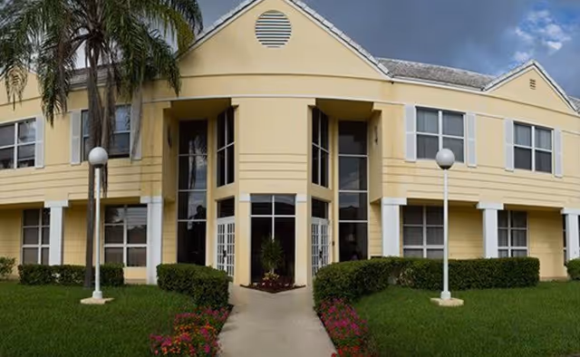 Yellow two-story senior living building with glass entrance, palm trees, and manicured lawn.