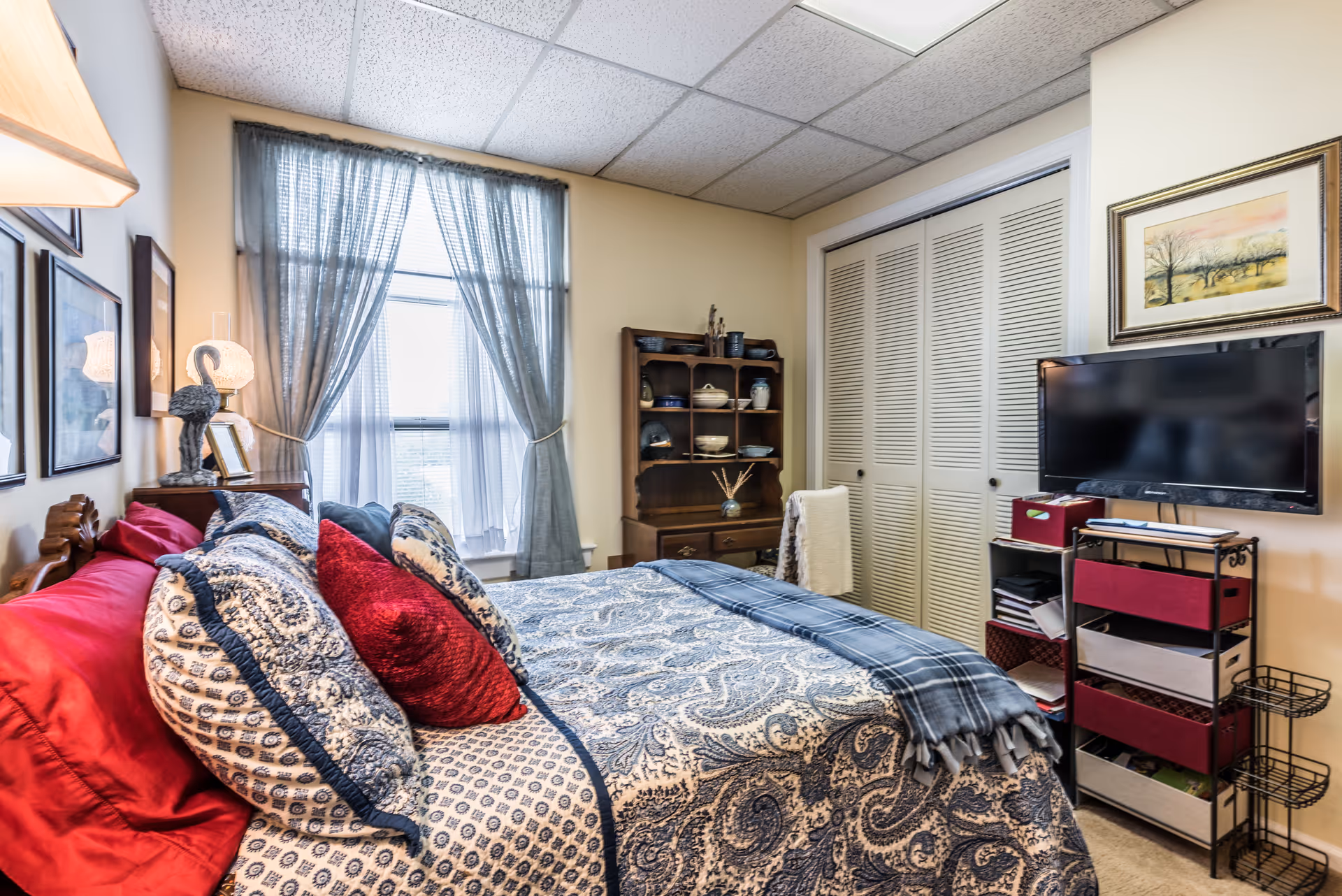 A cozy bedroom with a bed covered in patterned blue and white bedding and red accent pillows. The room has a window with sheer curtains letting in natural light, a wooden shelf with decorative items, a flat-screen TV mounted on the wall, and a closet with white louvered doors. There is a framed painting above the TV and a lamp on a bedside table.