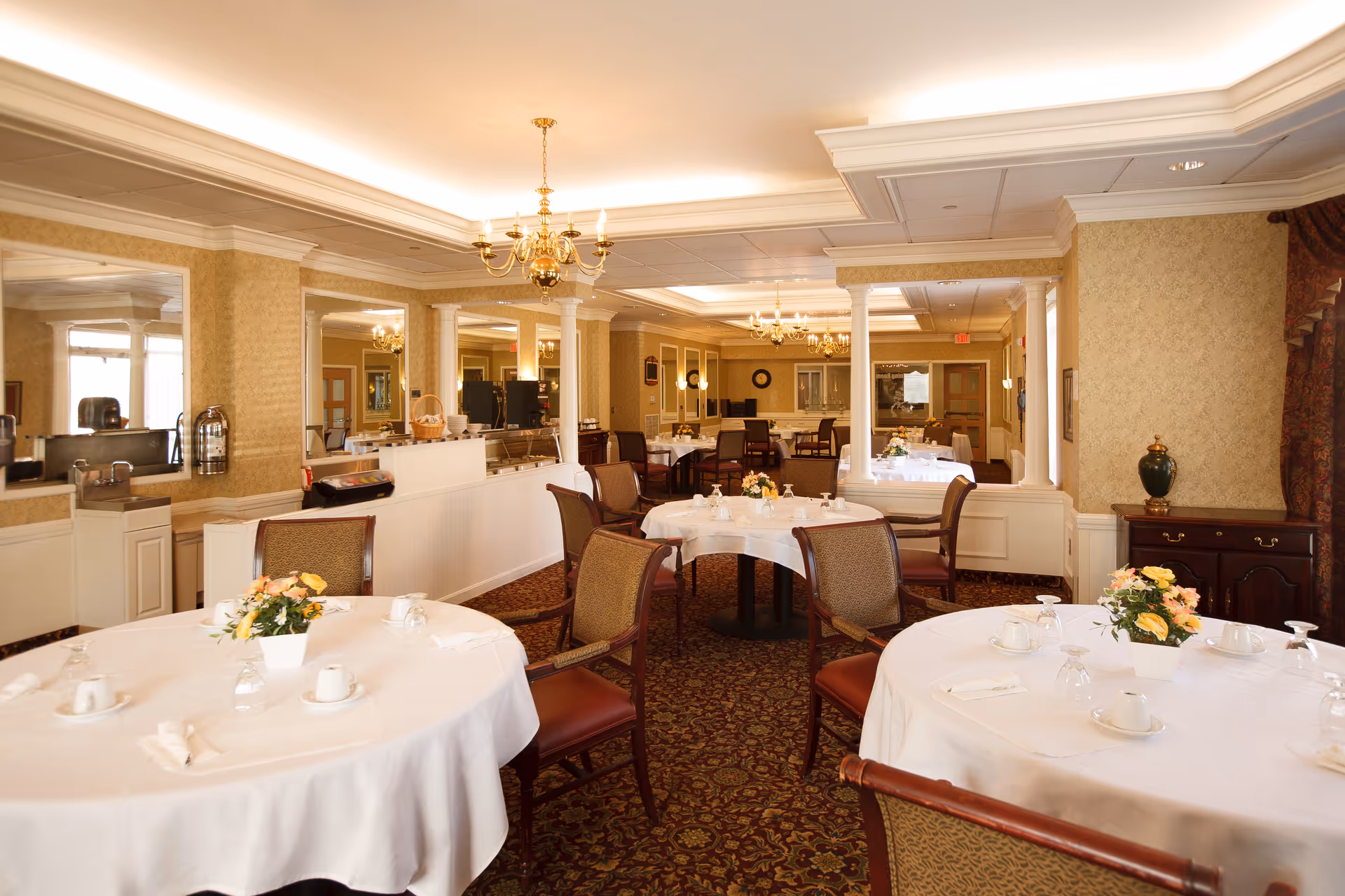 A well-lit dining room with round tables covered in white tablecloths, each set with cups, glasses, napkins, and floral centerpieces. The room features patterned carpet, upholstered chairs, chandeliers, and decorative columns. There is a serving area with coffee machines and mirrors on the walls.