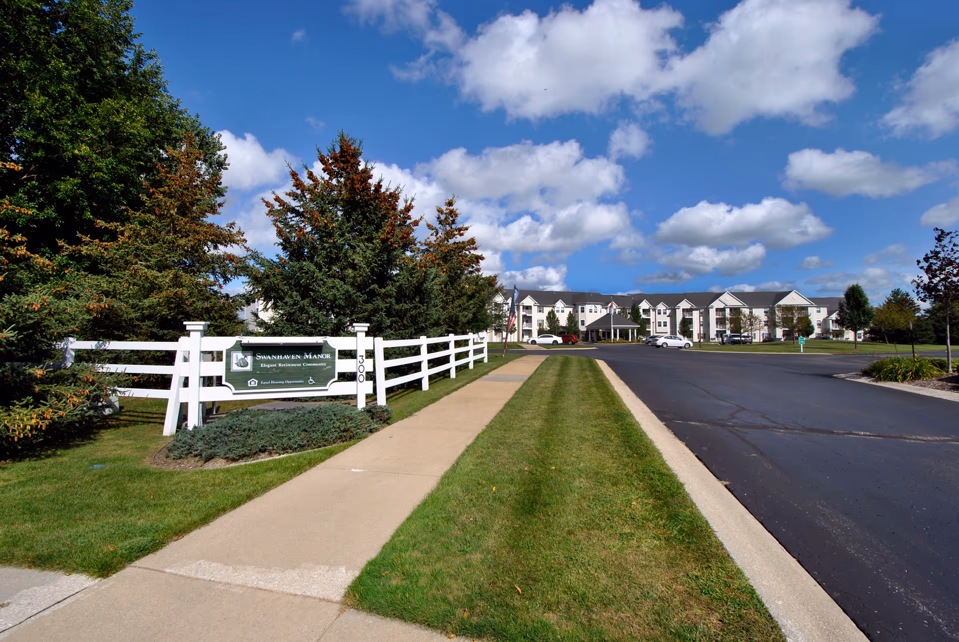 Sidewalk and driveway leading to the Swanhaven Manor retirement community building with a white fence and entrance sign under a blue sky.