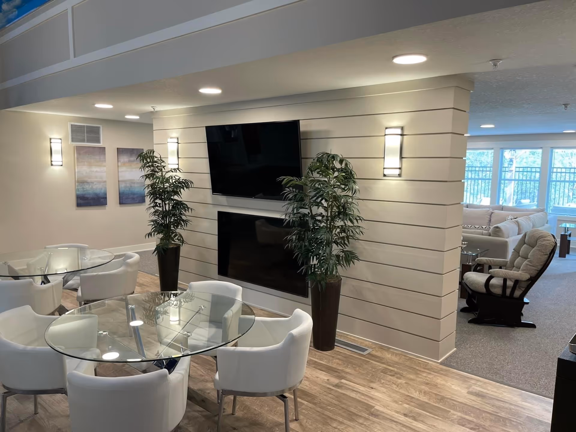 A modern interior common area featuring a glass round table surrounded by white cushioned chairs on a wooden floor. Two tall potted plants flank a wall-mounted flat-screen TV and a fireplace beneath it. In the background, there is a cozy seating area with a cushioned rocking chair and a large sectional sofa near windows letting in natural light.