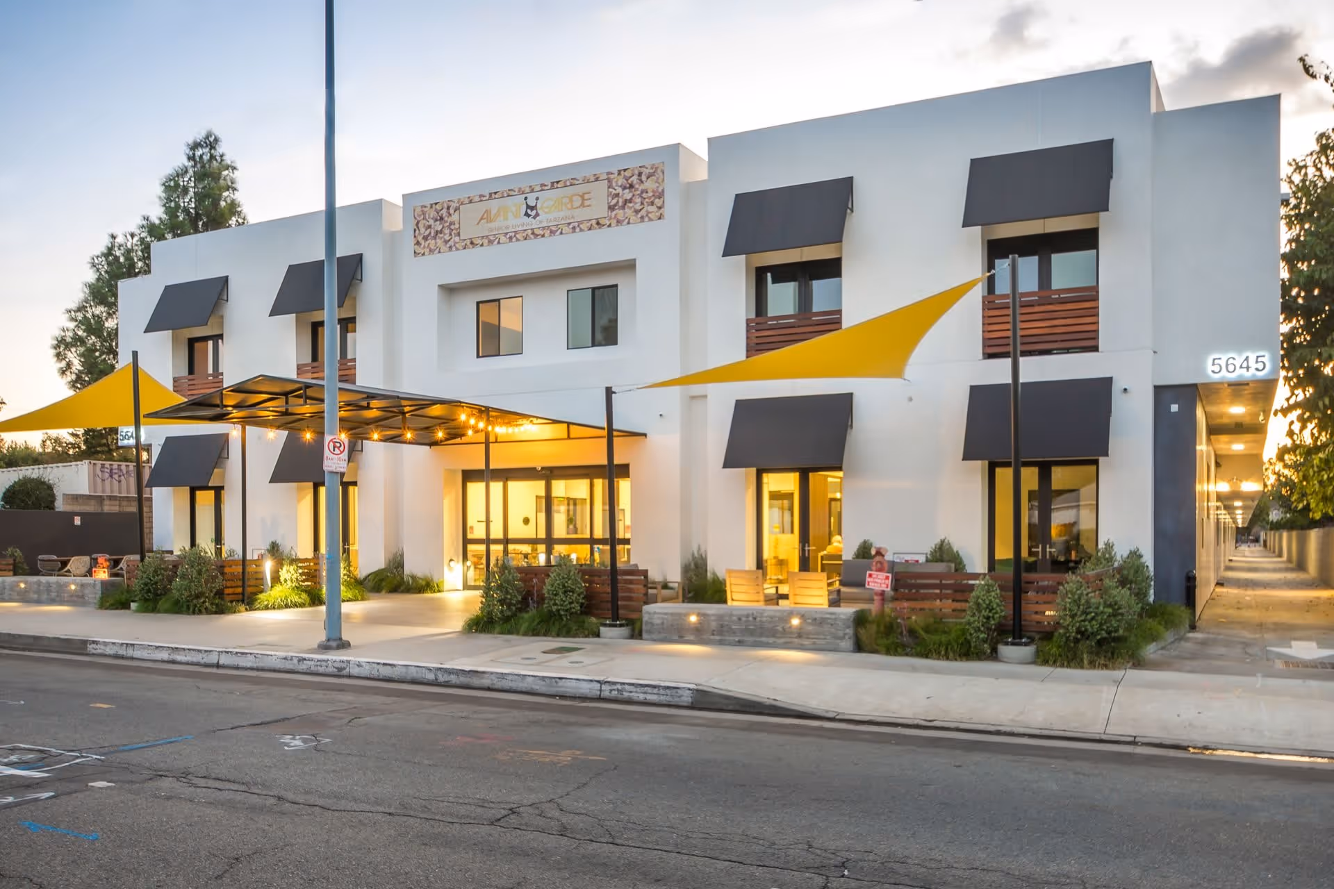 Front exterior of a modern two-story white senior living building labeled AvantGarde with yellow shade sails and a lit entrance.