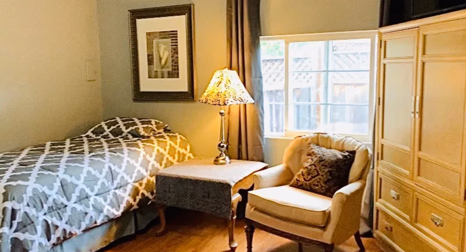 A cozy bedroom with a single bed covered in a gray and white patterned comforter, a wooden side table with a decorative lamp, a cushioned armchair with a patterned pillow, a window with curtains, and a wooden wardrobe.