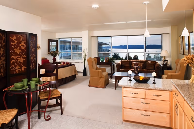 A spacious and well-lit living area in a retirement community apartment featuring a sitting area with a sofa, two armchairs, a coffee table, and a large window with a scenic view of water and distant hills. The room also includes a small dining table with chairs, a decorative folding screen, and a kitchen counter with cabinets and a bowl of fruit.