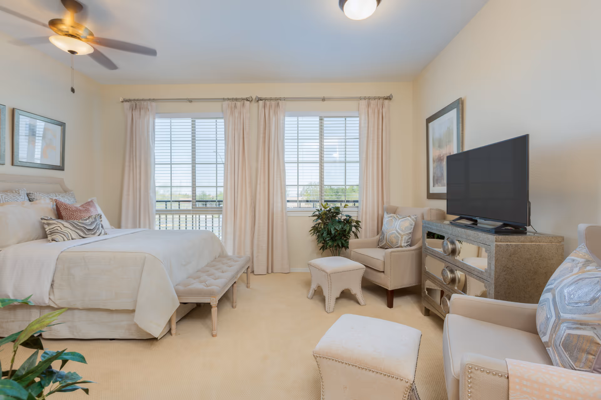 A bright and cozy bedroom with a large bed dressed in beige linens and multiple pillows. There is a cushioned bench at the foot of the bed, two beige armchairs with patterned pillows, a small matching ottoman, and a TV on a mirrored dresser. The room has large windows with beige curtains letting in natural light, a ceiling fan, and light-colored walls and carpet.