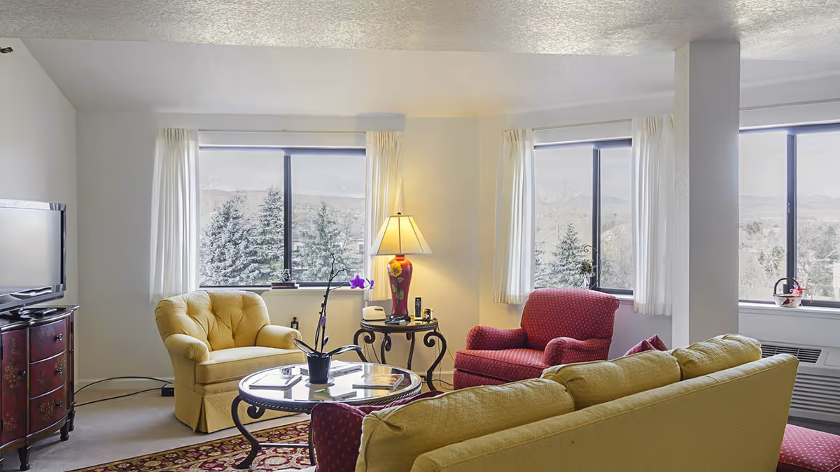 Bright living room with a yellow sofa, upholstered armchairs, coffee table and large windows overlooking trees and hills.