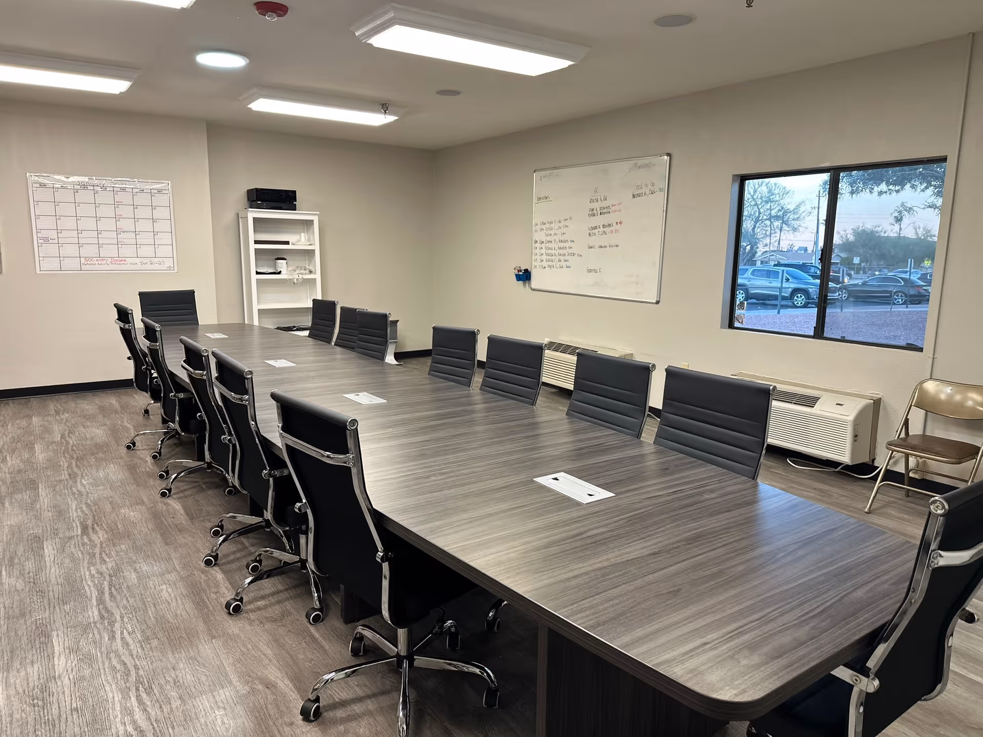 Long conference table with black rolling chairs in a meeting room featuring whiteboards, shelving, and a window showing parked cars.