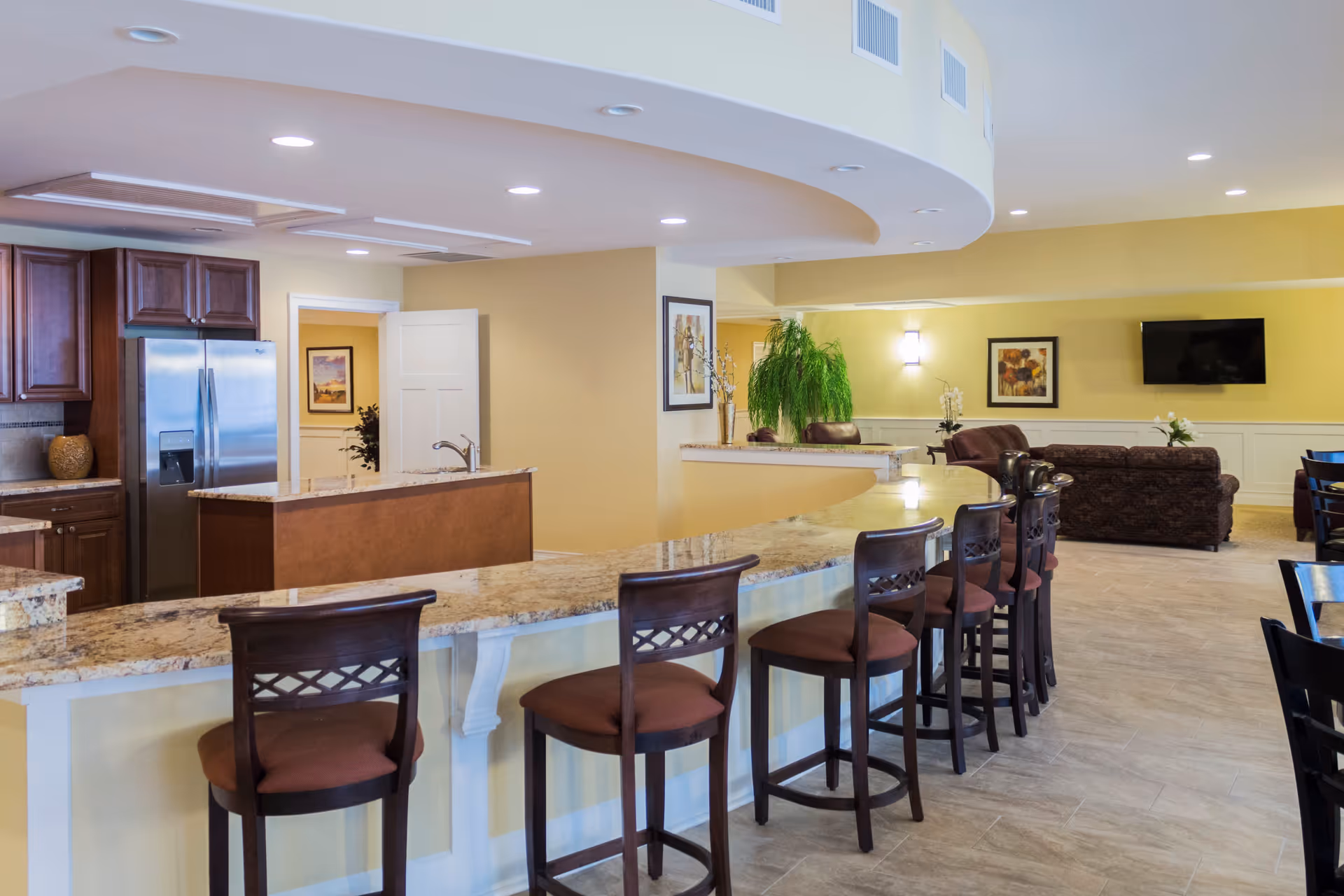 Interior view of a senior living facility featuring a long curved granite countertop with six wooden bar stools. Behind the counter is a kitchen area with wooden cabinets and a stainless steel refrigerator. The room extends into a living area with sofas, a wall-mounted TV, framed artwork, and a large green plant. The walls are painted light yellow, and the ceiling has recessed lighting.