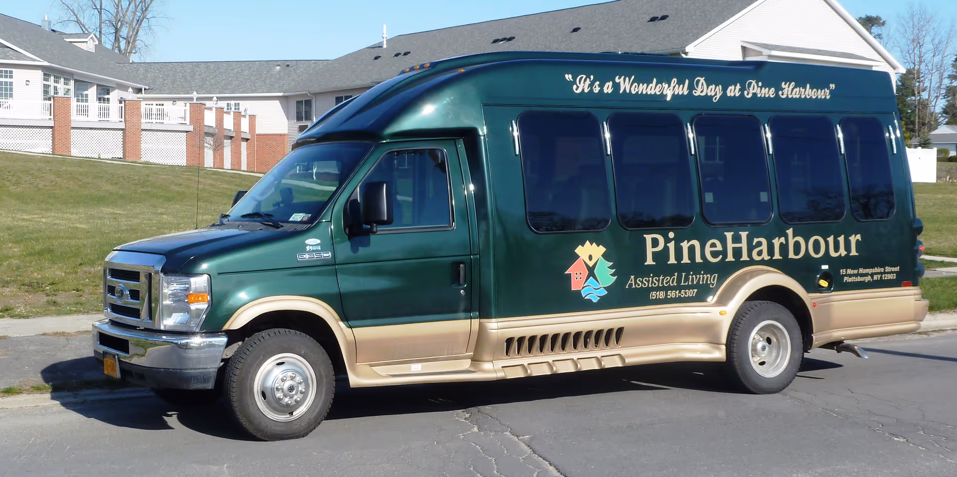 A green and beige shuttle van parked on a street in front of a residential building. The van has the text 'It's a Wonderful Day at Pine Harbour' and 'Pine Harbour Assisted Living' along with contact information and a logo on its side.