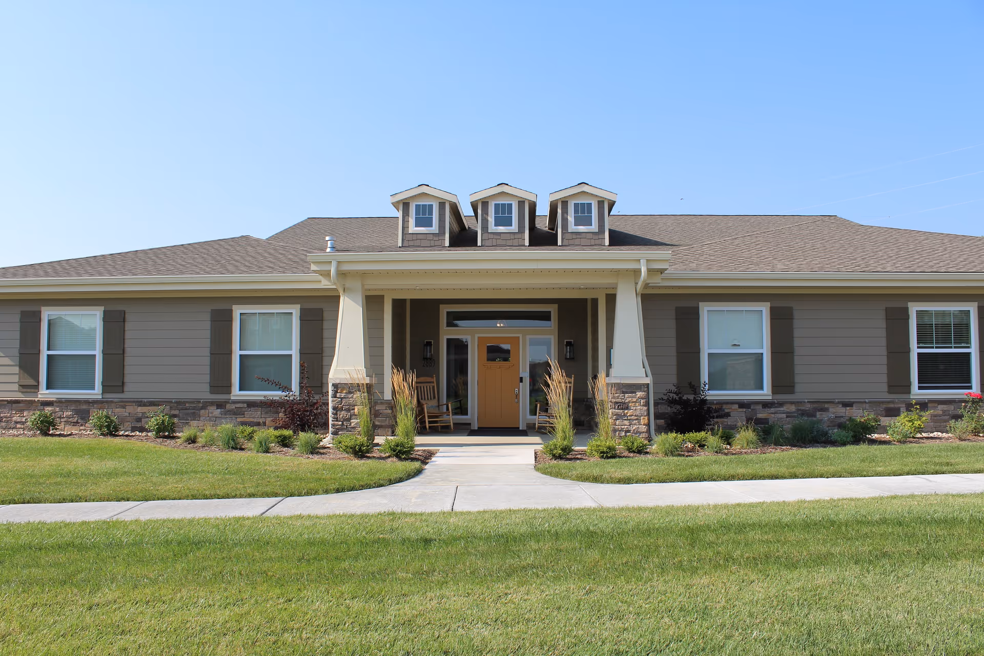 Front exterior view of a single-story building with a covered entrance supported by columns, three dormer windows on the roof, and a well-maintained lawn and landscaping in front.