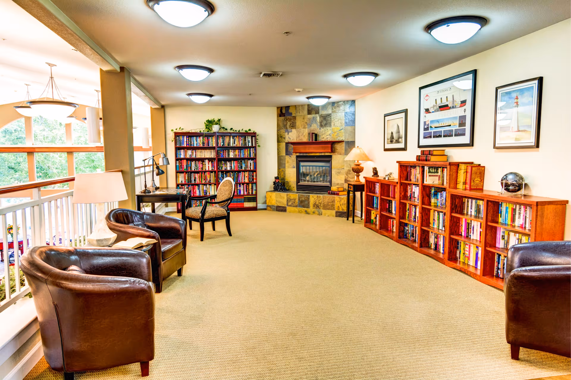 A cozy reading lounge area with leather armchairs, bookshelves filled with books, a stone fireplace, framed nautical-themed artwork on the walls, and soft overhead lighting.