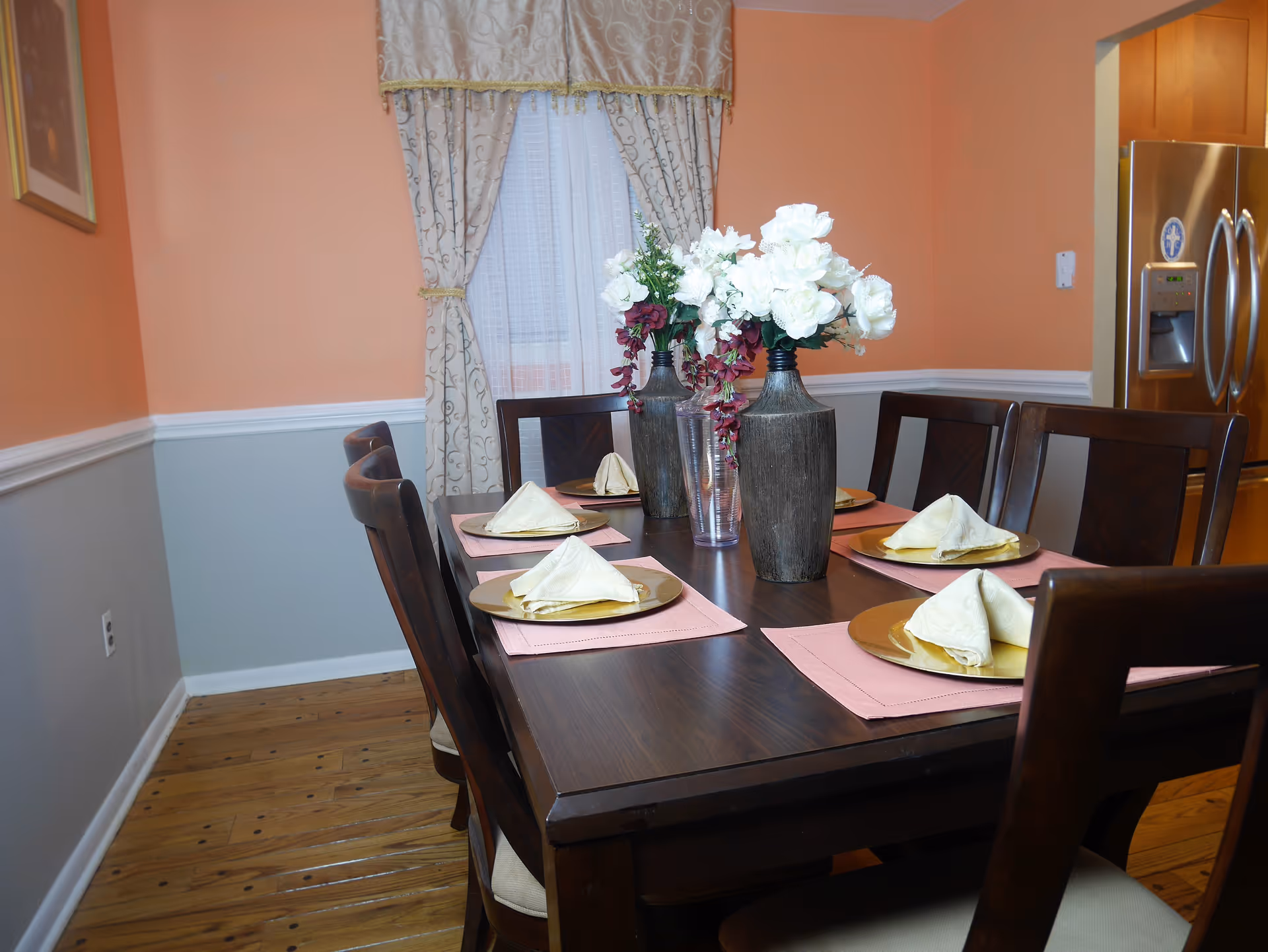 Dining room with a dark wood table set for six, floral centerpieces, chairs and a stainless refrigerator visible in the background.