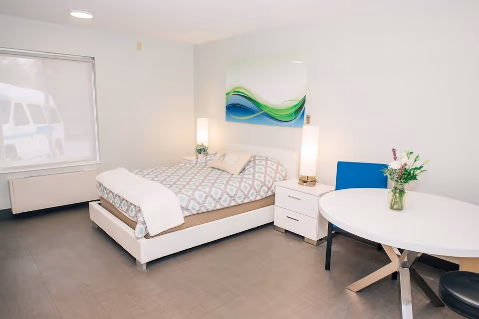 Bright modern bedroom with a bed, two nightstands and lamps, a round table with a vase of flowers, and abstract wall art.