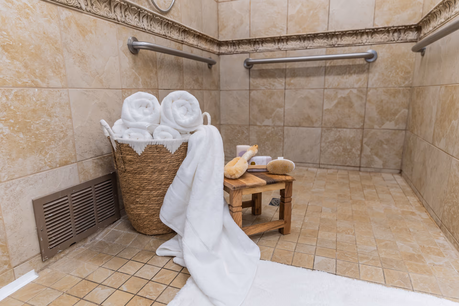 Tiled accessible shower with grab bars, a basket of rolled white towels and a small wooden stool holding bath brushes and a draped towel.