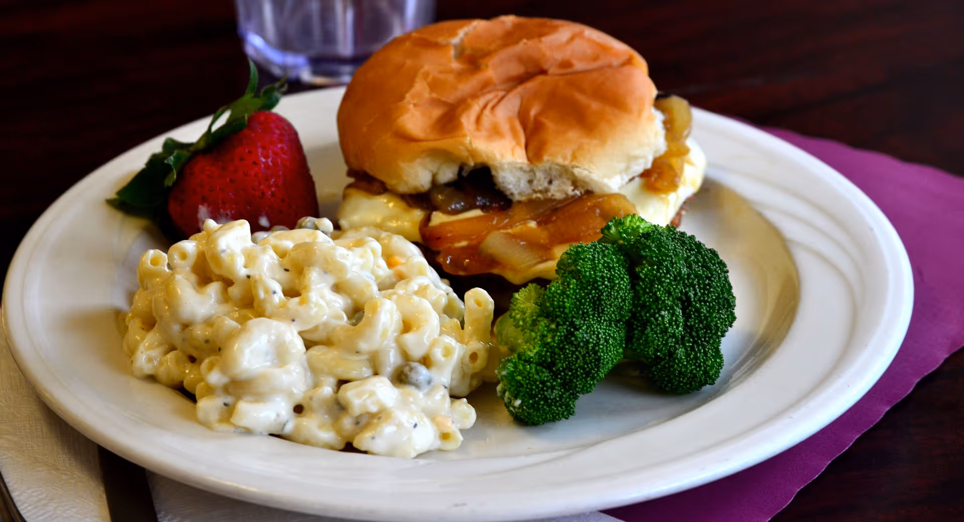 A plate with a cheeseburger sandwich, a serving of macaroni and cheese, a few pieces of broccoli, and a strawberry, placed on a table with a purple placemat and a glass of water in the background.