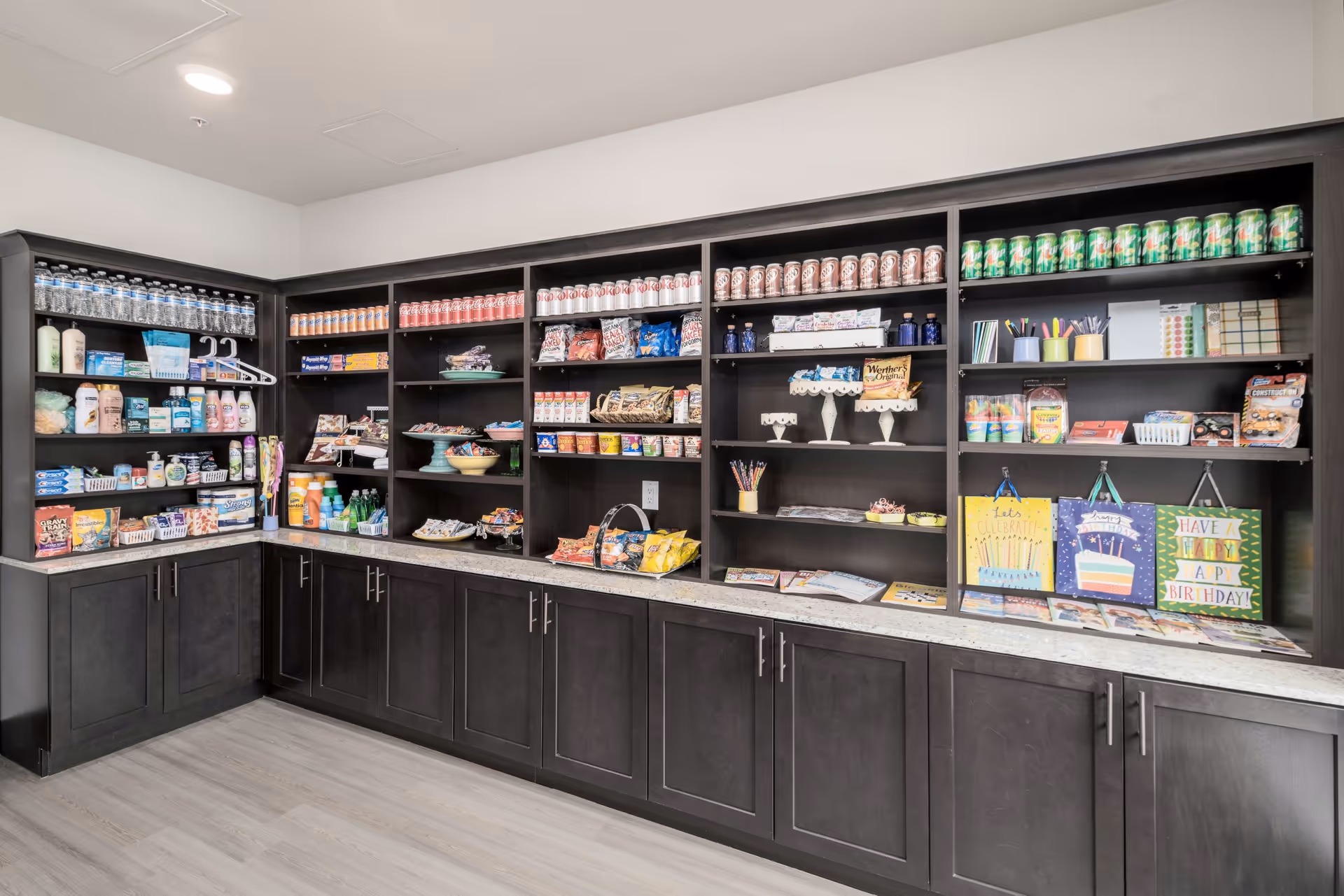 A corner shelving unit in a senior living facility stocked with various snacks, beverages, toiletries, and small gift items. The shelves contain bottled water, canned sodas, chips, candy, personal care products, and colorful gift bags. The cabinetry below is dark wood with silver handles, and the floor is light-colored wood.