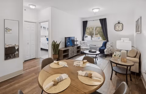 A bright and cozy living and dining area in a senior living facility. The foreground features a round wooden dining table set with four placemats and napkins. In the background, there is a living room with a TV on a wooden stand, two patterned armchairs, a small round coffee table, a beige sofa, and a side table with a lamp and decorative items. A window with dark curtains lets in natural light, and a wall clock is mounted above the sofa. The floor is wooden, and a doorway leads to a bedroom with a bed visible.