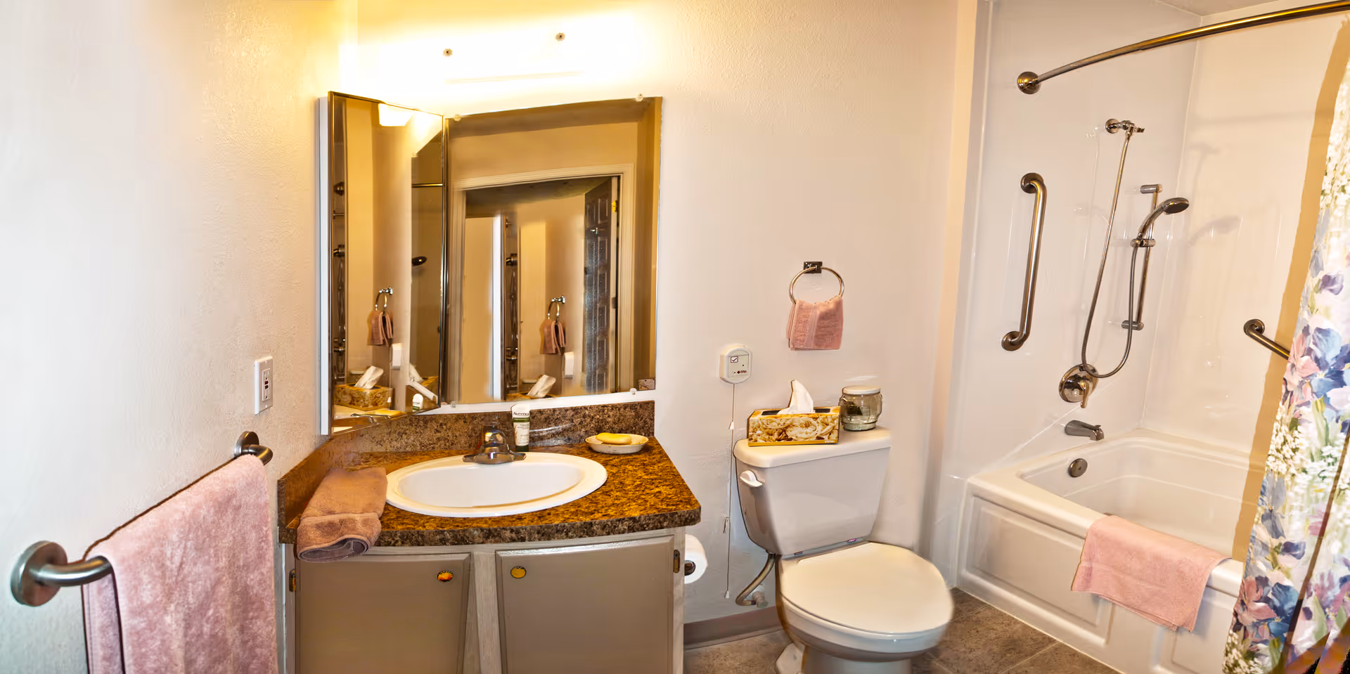 A bathroom with a sink and countertop on the left, a toilet in the center, and a bathtub with a shower on the right. The countertop has a brown towel, a soap dish, and a small bottle. The toilet tank has a tissue box and a candle. The bathtub has grab bars and a pink towel hanging on the side. A floral shower curtain is partially visible on the right side. The walls are light-colored, and there is a large mirror above the sink.