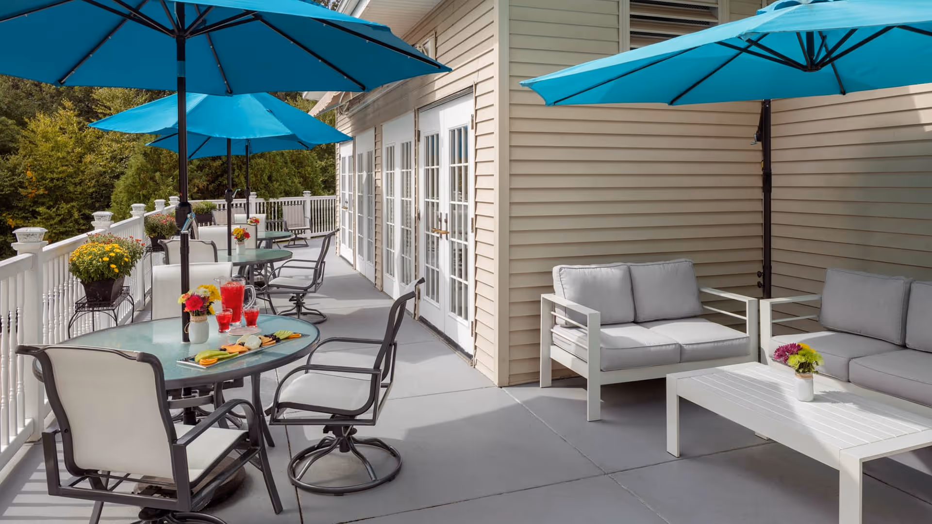 Outdoor patio area with several glass-top tables and chairs under blue umbrellas. There are also white cushioned sofas and a white coffee table with a small flower arrangement. The patio is adjacent to a beige building with multiple glass doors and surrounded by greenery.