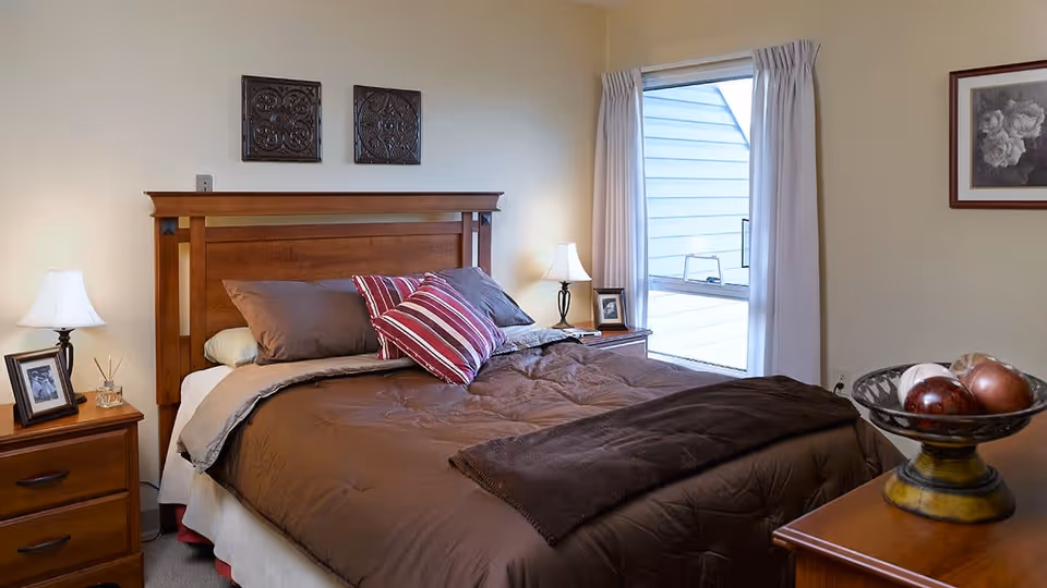 A cozy bedroom with a wooden bed frame and headboard, made up with brown bedding and pillows including a striped accent pillow. There are two wooden nightstands on either side of the bed, each with a lamp and framed photo. A window with white curtains is on the right wall, and a decorative bowl with ornamental balls sits on a wooden dresser in the foreground. The walls are painted a light beige color with framed artwork hanging.