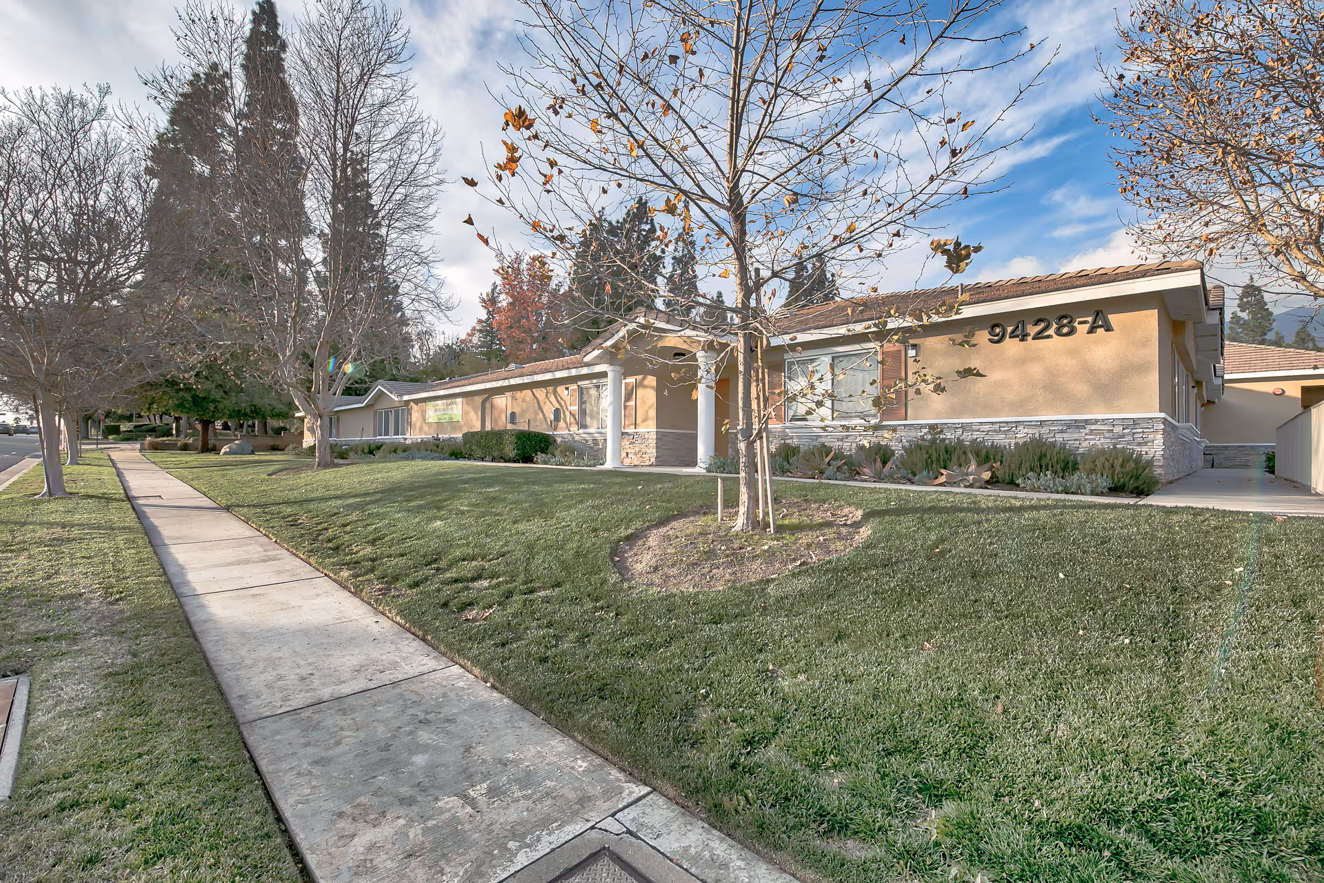 Exterior view of a single-story assisted living facility building with the address number 9428-A displayed on the wall. The building is surrounded by a well-maintained lawn, leafless trees, and a sidewalk running alongside the property under a partly cloudy sky.
