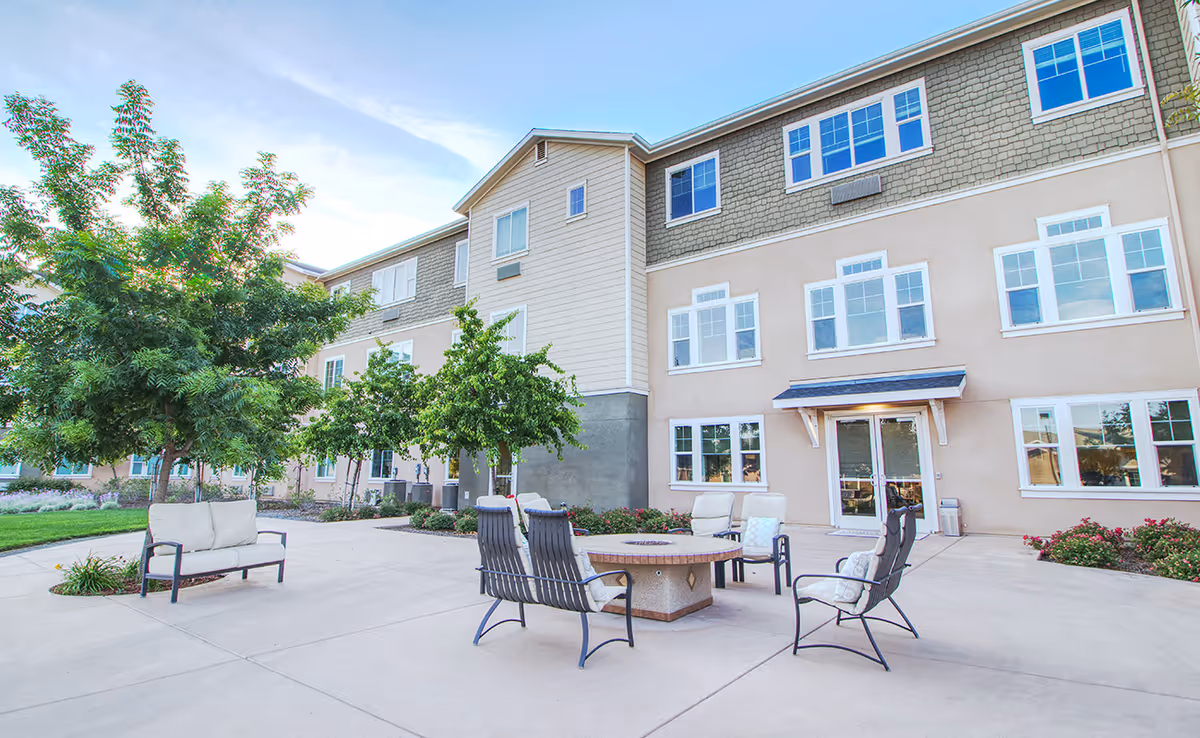 Outdoor patio area at Carmel Village Memory Care with cushioned chairs and a circular fire pit table, surrounded by trees and landscaping, adjacent to a three-story building with multiple windows and a double glass door entrance.
