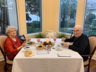 An elderly woman and an elderly man sitting at a dining table set with plates of food, drinks, and a centerpiece. They are in a room with large windows showing trees and greenery outside.
